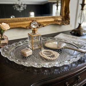 May include: A decorative glass tray holds a perfume bottle, a hand mirror, a pearl necklace, and a small ring box. A small vase with a rose and a gold-framed mirror are in the background. The tray is on a dark wooden surface.