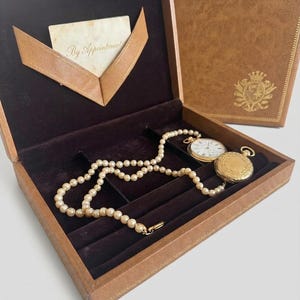 May include: Open brown leather jewelry box with a pearl necklace, a gold pocket watch, and a second closed box. The open box has a dark brown velvet interior and a card that reads "By Appointment". The closed box has a gold emblem.