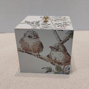 May include: A white box with a gold knob on the top. The box has a floral design with two birds on a branch. The birds are brown and white.