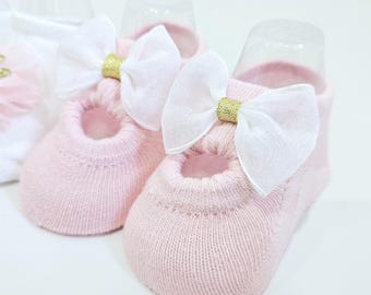 Könnte beinhalten: Ein Paar rosa Babysocken mit weißen Schleifen und goldenen Akzenten. Die Socken haben einen zarten, spitzigen Look und sind perfekt für einen besonderen Anlass.
