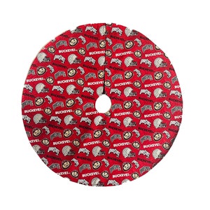 May include: Red Christmas tree skirt with a repeating pattern of football helmets, the word "BUCKEYES", and the Ohio State University logo. The skirt has a white center hole and is circular.
