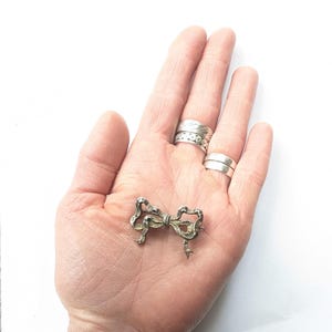 May include: A silver-toned bow brooch with intricate detailing. The brooch is in the shape of a tied bow, with delicate loops and tails. The brooch is displayed on a hand, with several silver rings on the fingers.