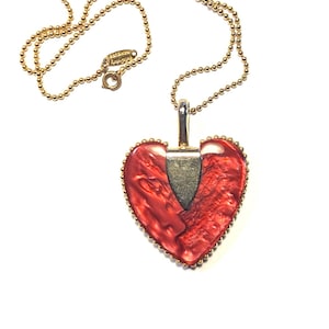 Puede incluir: Un collar dorado con un colgante en forma de corazón. El colgante presenta un diseño rojo veteado con un borde dorado y un acento plateado. La cadena del collar es una delicada cadena de bolas.