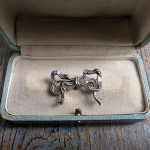 May include: A silver-toned bow brooch displayed inside a vintage, light blue jewelry box. The brooch is intricately detailed, with a textured surface. The box has a light beige interior and a gold-colored clasp.