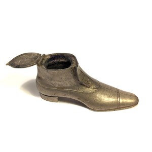 May include: A silver metal shoe-shaped trinket box with a hinged lid. The box is detailed with a decorative design and has a small heel.