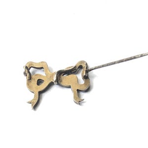 May include: A vintage gold-toned bow pin with a long, silver-colored pin extending from the back. The bow has a detailed, ornate design with curved lines and small loops. The pin is set against a white background.