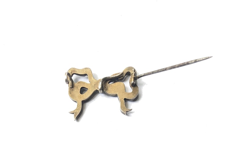 May include: A vintage gold-toned bow pin with a long, silver-colored pin extending from the back. The bow has a detailed, ornate design with curved lines and small loops. The pin is set against a white background.
