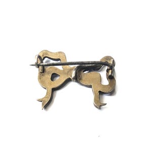 May include: A vintage bow-shaped brooch in a gold tone, with a silver-colored pin. The brooch has a detailed design with curved lines and a classic bow shape. The pin is attached to the back of the brooch.