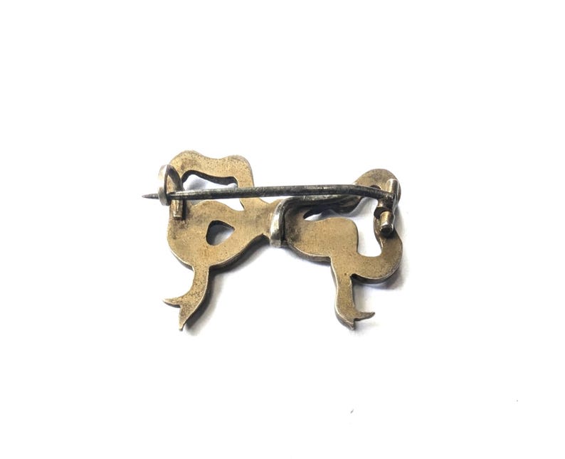 May include: A vintage bow-shaped brooch in a gold tone, with a silver-colored pin. The brooch has a detailed design with curved lines and a classic bow shape. The pin is attached to the back of the brooch.