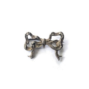 May include: A silver-toned bow brooch with a textured surface. The brooch is in the shape of a tied bow, with detailed loops and tails. The pin is attached to the back of the bow.