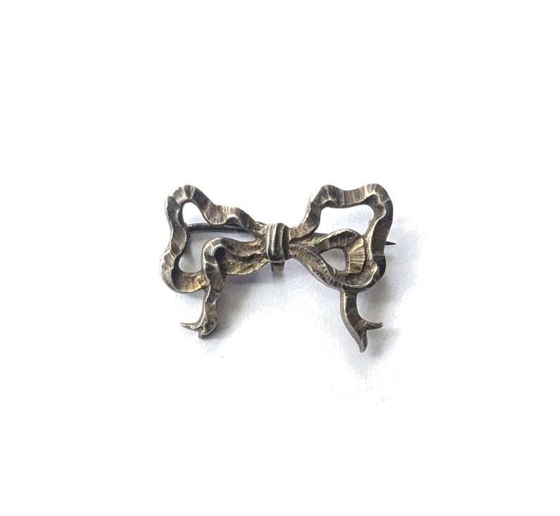 May include: A silver-toned bow brooch with a textured surface. The brooch is in the shape of a tied bow, with detailed loops and tails. The pin is attached to the back of the bow.