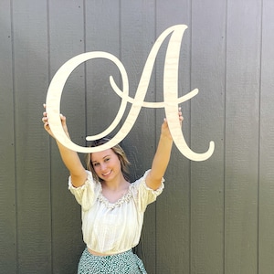 May include: A person holds up a large, wooden, cursive letter A. The letter is painted white and has a light wood grain texture.