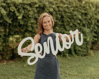 Peut inclure: Une enseigne en bois découpée de couleur claire avec le mot Senior écrit en lettres cursives. L'objet est tenu horizontalement et présente une typographie fluide aux bords arrondis avec une finition mate, servant d'accessoire décoratif.
