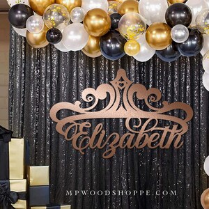 May include: A gold and black party backdrop with a custom wooden sign that says "Elizabeth" in a decorative font. The sign is shaped like a crown and is surrounded by a garland of gold and black balloons.