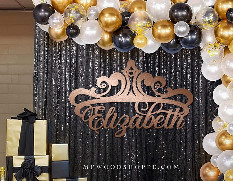 May include: A gold and black party backdrop with a custom wooden sign that says "Elizabeth" in a decorative font. The sign is shaped like a crown and is surrounded by a garland of gold and black balloons.