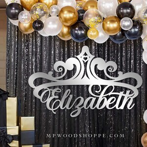May include: Silver glitter backdrop with a silver crown and the name "Elizabeth" cut out in a decorative font. The backdrop is decorated with black, gold, and white balloons.
