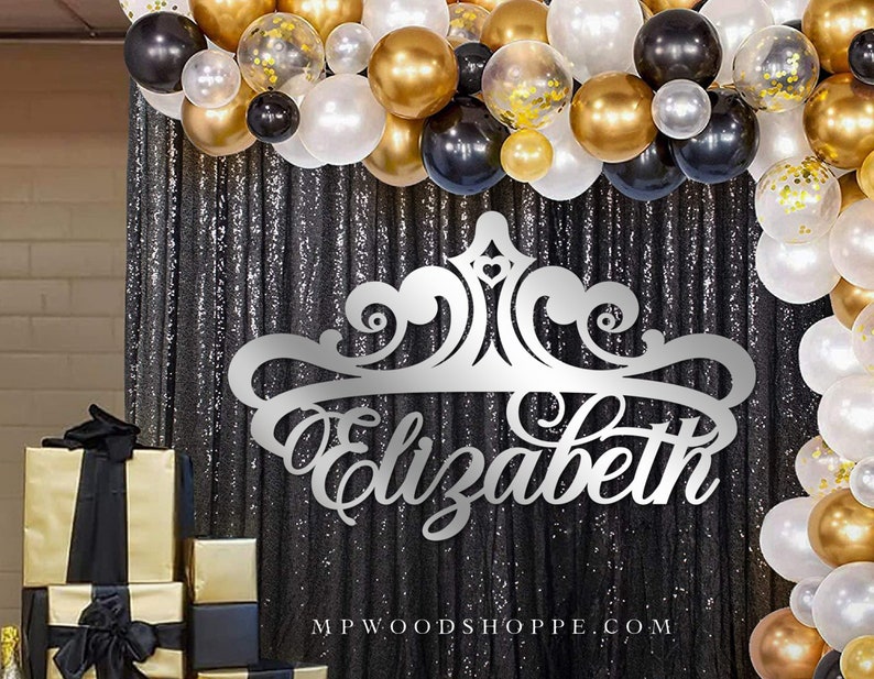 May include: Silver glitter backdrop with a silver crown and the name "Elizabeth" cut out in a decorative font. The backdrop is decorated with black, gold, and white balloons.