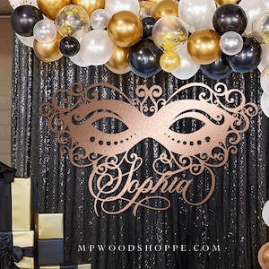 K&ouml;nnte beinhalten: Eine ros&eacute;goldene Maskenballmaske mit filigranen Details und dem Namen "Sophia" in dekorativer Schrift. Die Maske wird vor einem schwarzen Paillettenhintergrund mit einem Ballonbogen aus schwarzen, goldenen und wei&szlig;en Ballons pr&auml;sentiert.