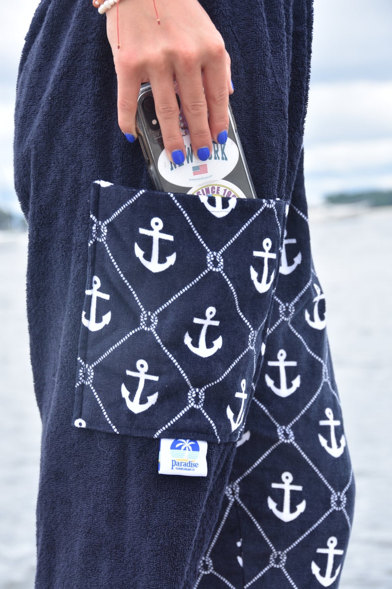 May include: A navy blue towel with a large pocket featuring a white anchor pattern. The pocket has a smaller label with the text "Paradise" and a palm tree logo. The towel has a white label with the text "Since 1982".