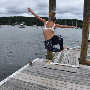 May include: A woman in a white bikini top and blue and white anchor print pants jumps off a wooden dock into the water. The dock is over a body of water with boats in the distance.
