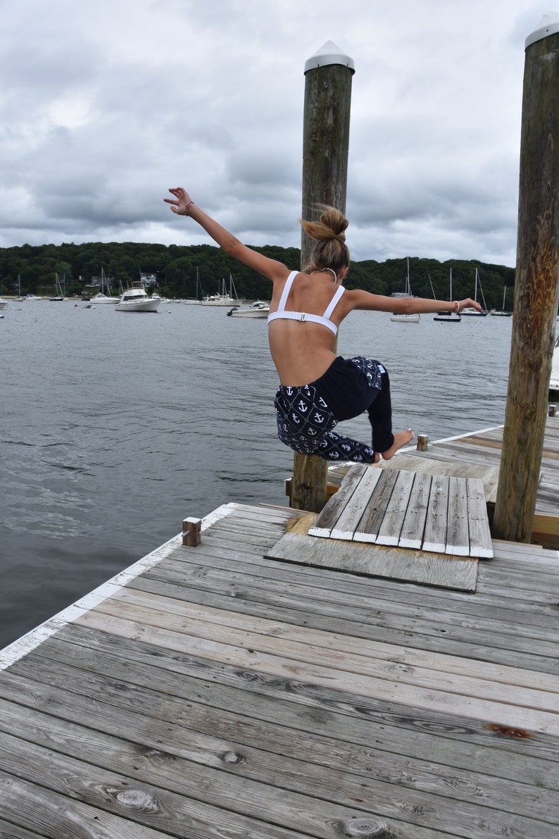 May include: A woman in a white bikini top and blue and white anchor print pants jumps off a wooden dock into the water. The dock is over a body of water with boats in the distance.