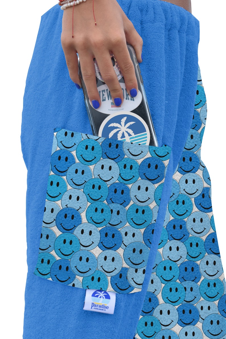 May include: Blue terry cloth shorts with a large pocket on the side. The pocket is lined with a white background and a pattern of blue smiley faces. The shorts have a white label with the text "Paradise" and a palm tree logo.