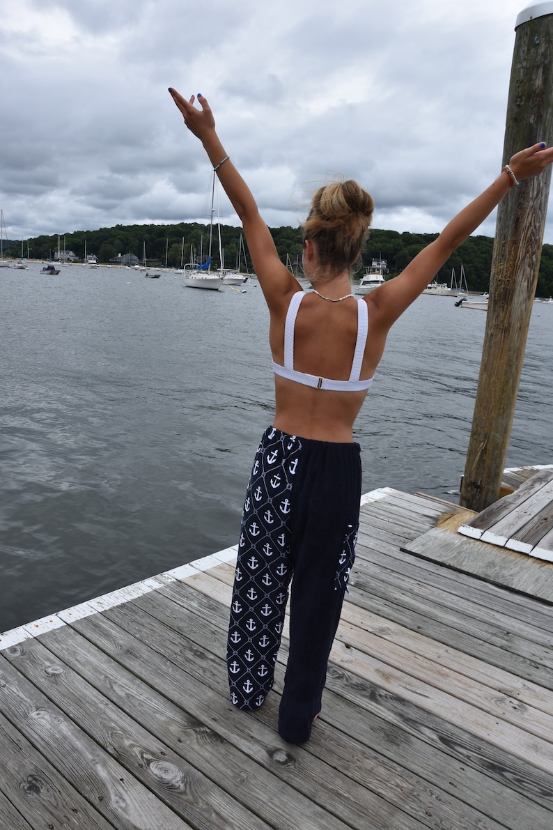 May include: A woman wearing a white halter top and navy blue pants with a white anchor pattern stands on a wooden dock with her arms raised above her head. The water is in the background.