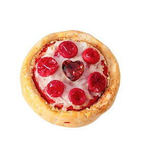 May include: A round, textured beige crust surrounds a red centre topped with white icing, five small red circular accents, and a single central dark red heart-shaped gem. The overall appearance resembles a small, decorative pastry or tart.