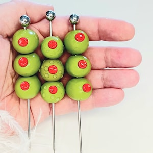 Puede incluir: Tres pinchos de aceitunas con tres aceitunas verdes en cada pincho. Las aceitunas tienen centros rojos y están dispuestas en fila en una mano.