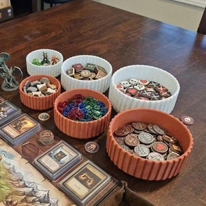 May include: A collection of round, ribbed storage bowls in white and terracotta colours, filled with various board game components including small coloured cubes, circular tokens with skull and heart icons, and miniature game pieces.