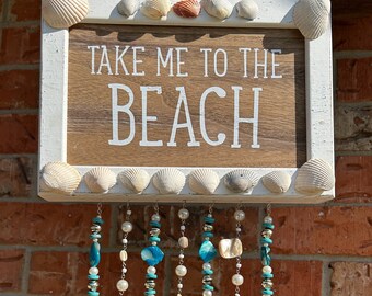 May include: A white wooden sign with the text "Take Me To The Beach" surrounded by seashells and hanging blue and white beaded strands with seashells and starfish.