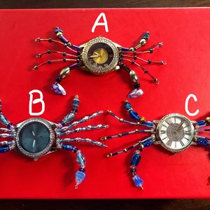 May include: Three jeweled crab-shaped wristwatches with different colored faces. The top watch has a gold face with black numbers, the middle watch has a blue face with silver hands, and the bottom watch has a white face with black Roman numerals.