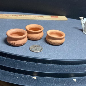 May include: Three small terracotta pots, each with a slightly different shape, are arranged on a dark gray surface. A silver coin is placed between two of the pots.