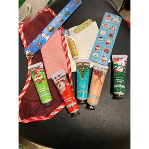 Puede incluir: Un conjunto de seis pequeñas cremas de manos con diseños navideños. Las cremas están en tubos de colores verde, azul, rojo y blanco. Los tubos tienen las palabras "Feliz Navidad" impresas. También hay un pequeño bálsamo labial con un diseño rojo y blanco y las palabras "Bálsamo labial" impresas.