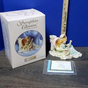 Puede incluir: Una figurita de porcelana blanca de dos ángeles tocando música. Los ángeles están sentados en una nube y están rodeados de flores. La figurita está en su caja original, que es blanca con un diseño rosa y azul. La caja también incluye un certificado de autenticidad.