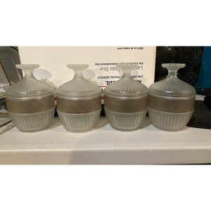 Puede incluir: Conjunto de cuatro tarros de vidrio esmerilado con ribetes dorados y un diseño decorativo. Los tarros son redondos y tienen una tapa.