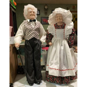 Puede incluir: Un par de muñecas de estilo vintage vestidas con atuendos formales. El muñeco masculino lleva una camisa blanca, un chaleco negro con un patrón blanco y negro, pantalones negros y zapatos negros. Lleva un bastón negro. La muñeca femenina lleva un vestido blanco con ribete rojo, un delantal blanco y un gorro blanco. Está de pie sobre una base con un patrón floral.