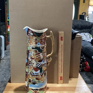 Op de afbeelding: Een hoge, keramische kan met een gouden handvat. De kan is versierd met een kleurrijk Japans geïnspireerd ontwerp met figuren en bloemen. De kan staat op een houten tafel.