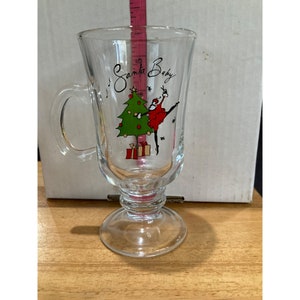 Puede incluir: Una taza de vidrio transparente con asa, con un diseño navideño. El diseño incluye un árbol de Navidad verde con regalos rojos y dorados, y una figura de Papá Noel vestido de rojo bailando con una media roja y blanca a rayas. El texto "Santa Baby" está escrito en cursiva en la taza.
