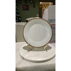 Puede incluir: Plato de cena de vidrio de leche blanco con un borde dorado y un patrón de remolino. El plato está en un soporte blanco.