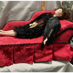 May include: A porcelain doll dressed in a black lace dress and a black shawl with fringe, lying on a red velvet chaise lounge with black trim.