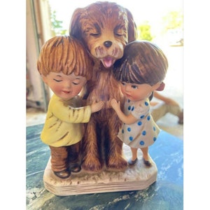 Op de afbeelding: Een keramieken beeldje dat een bruine hond afbeeldt die door twee kinderen wordt omhelsd. De kinderen, een in een geel shirt en de ander in een witte jurk met blauwe stippen, hebben hun ogen gesloten. De hond steekt zijn tong uit.