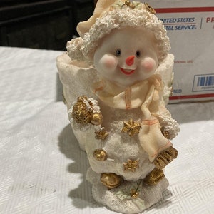 Könnte beinhalten: Weiße Keramik-Schneemann-Figur mit goldener Glitzer-Oberfläche. Der Schneemann trägt einen weißen Hut mit goldenen Verzierungen und einen cremefarbenen Schal mit goldenen Schneeflocken. Der Schneemann hat eine rote Nase und schwarze Augen.