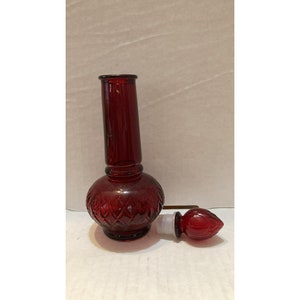 Könnte beinhalten: Eine rote Glasflasche mit einem Diamantenmuster und einem passenden roten Glasstopfen. Die Flasche hat einen hohen, schlanken Hals und einen runden Körper.