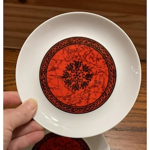 Puede incluir: Un platillo de cerámica blanco con un diseño rojo y negro. El diseño presenta un fondo rojo marmoleado con un patrón floral negro y un borde negro.