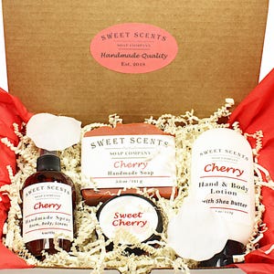 May include: A gift box filled with cherry-scented bath and body products from Sweet Scents Soap Company. The box contains a bar of soap, a bottle of hand and body lotion, a bottle of room spray, and a small jar of body butter. All products are labeled with the scent "Cherry" and the company name.