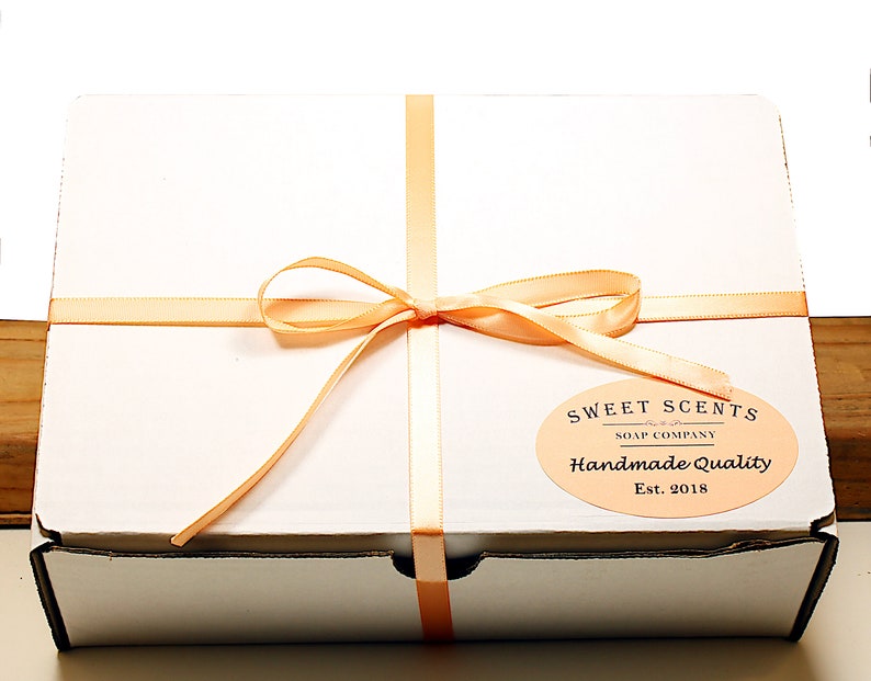 May include: A white gift box with a peach-colored ribbon tied in a bow. The box has a label that reads "Sweet Scents Soap Company Handmade Quality Est. 2018."