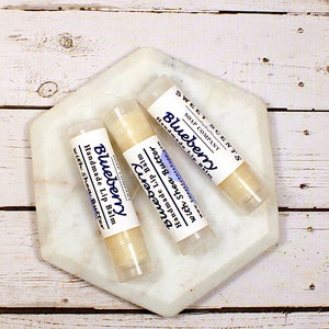 May include: Three tubes of handmade lip balm with the text "Blueberry" and "with Shea Butter" on a white marble hexagon. The lip balm tubes are labelled "Sweet Scents Soap Company". The background is a white wooden surface.