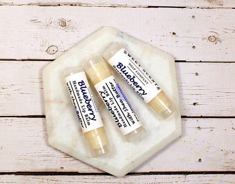 May include: Three tubes of handmade lip balm with the text "Blueberry" and "with Shea Butter" on a white marble hexagon. The lip balm tubes are labelled "Sweet Scents Soap Company". The background is a white wooden surface.