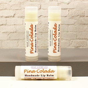 K&ouml;nnte beinhalten: Drei wei&szlig;e Lippenbalsam-Tuben mit einem wei&szlig;en Etikett, auf dem "SOAP COMPANY" und "Pina Colada Handmade Lip Balm" in orangefarbenem und schwarzem Text steht.